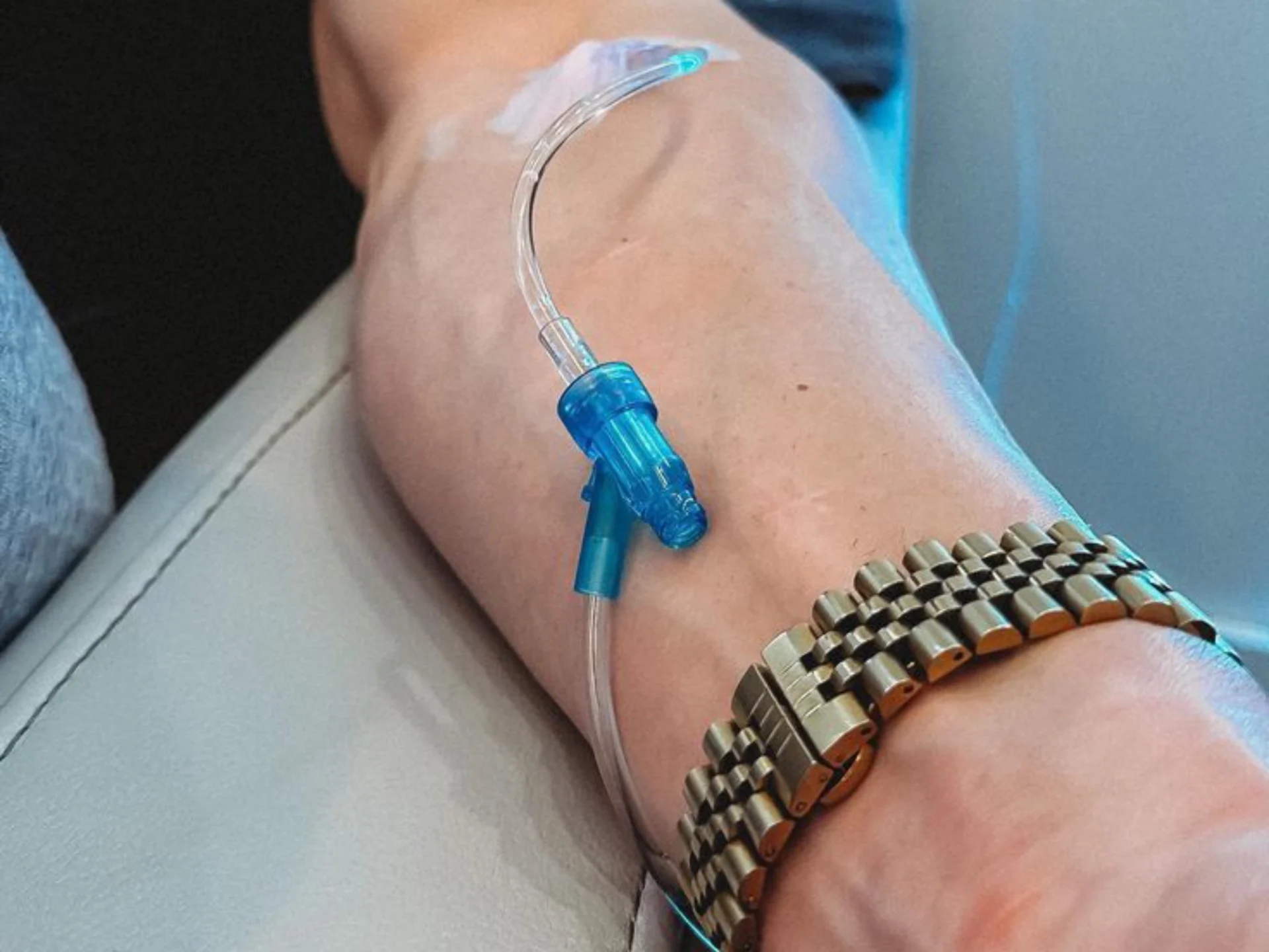 Close-up of a medical IV line taped to the neck/upper chest area, with a blue connector and clear tubing, next to a metal watch on the wrist