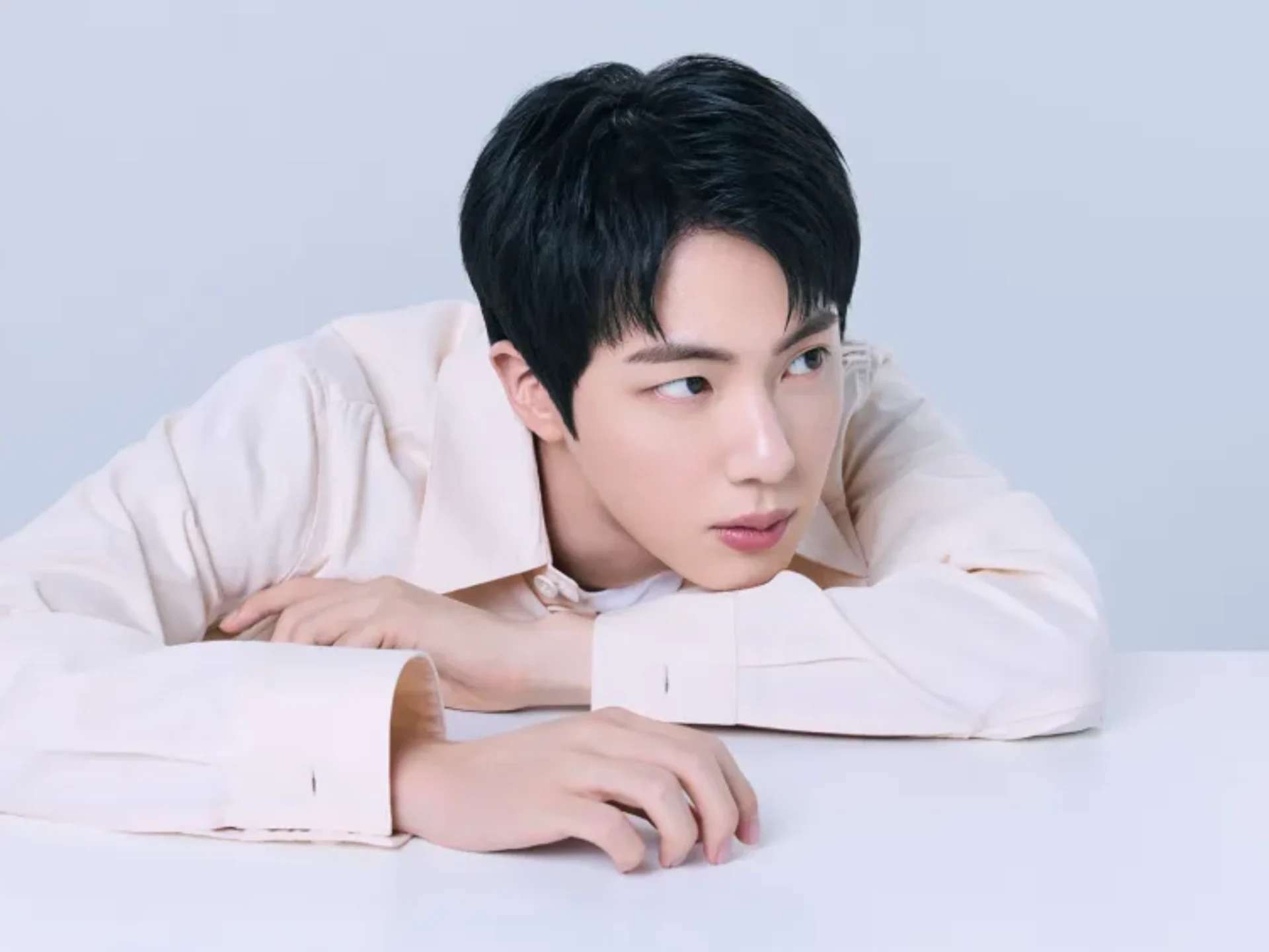 Young man with short black hair rests his chin on folded arms on a white desk, looking down at papers in front of him.