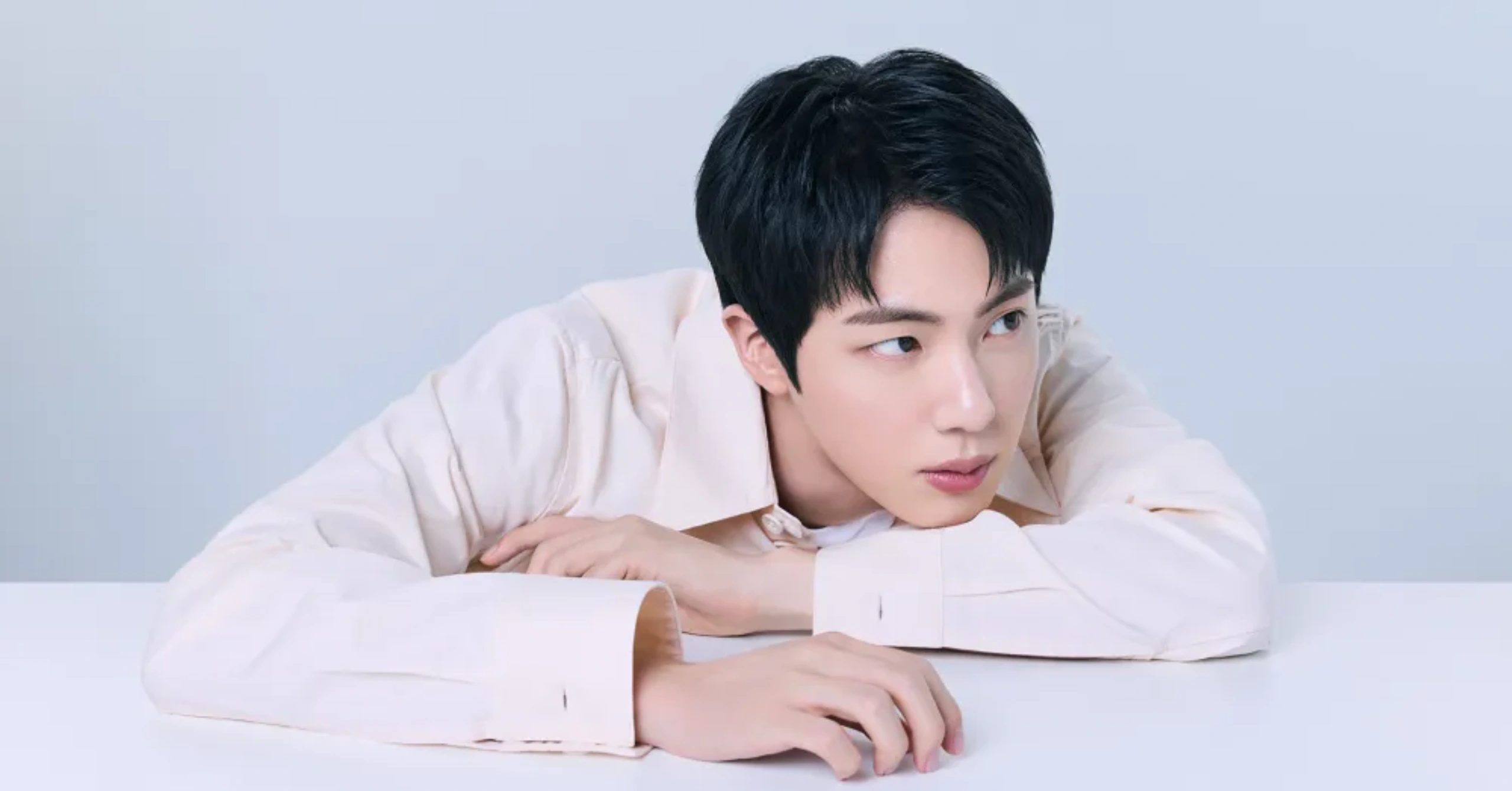 Young man with short black hair rests his chin on folded arms on a white desk, looking down at papers in front of him.