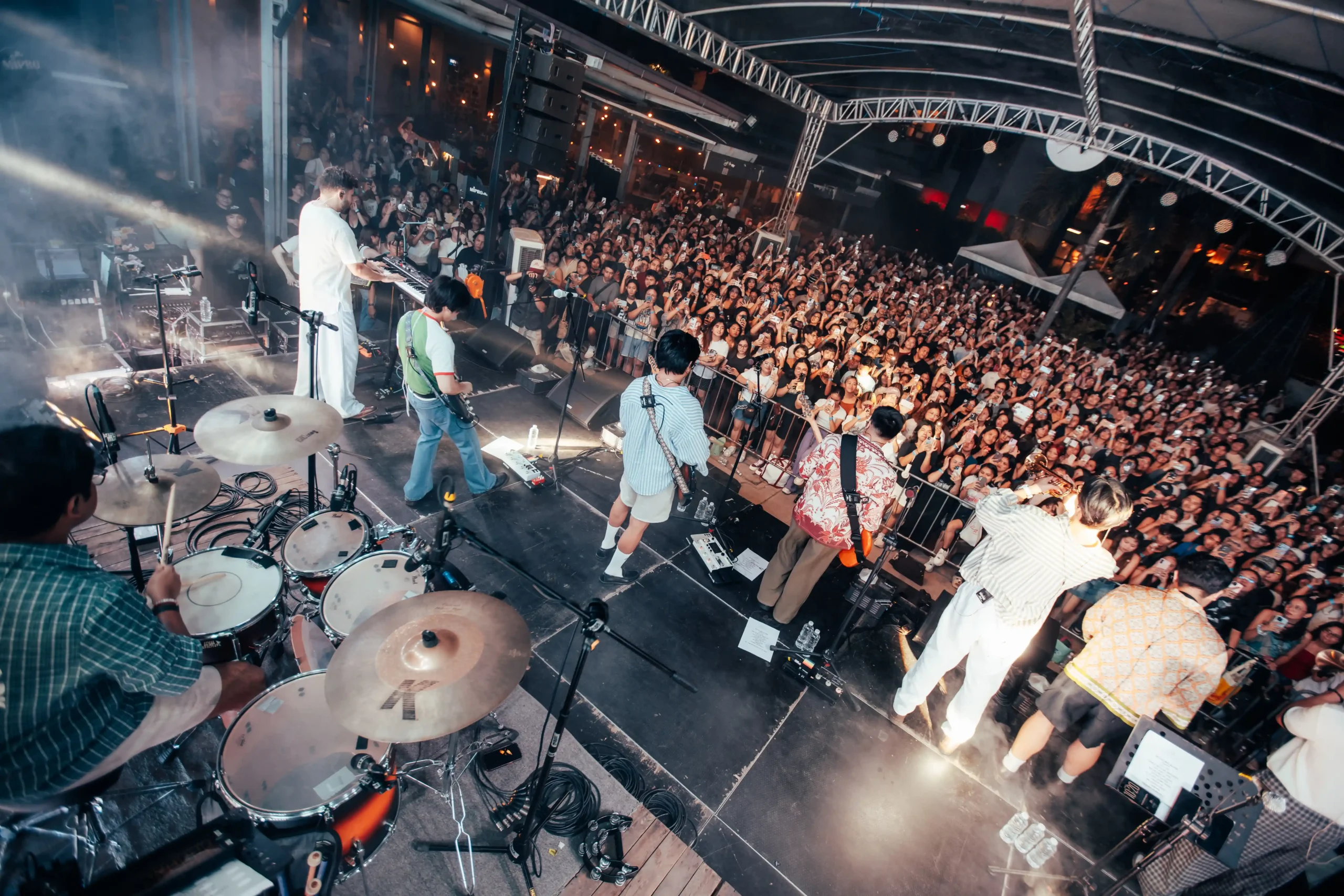 Live outdoor concert on a stage with a drummer in the foreground and a dense cheering crowd under metal trusswork.