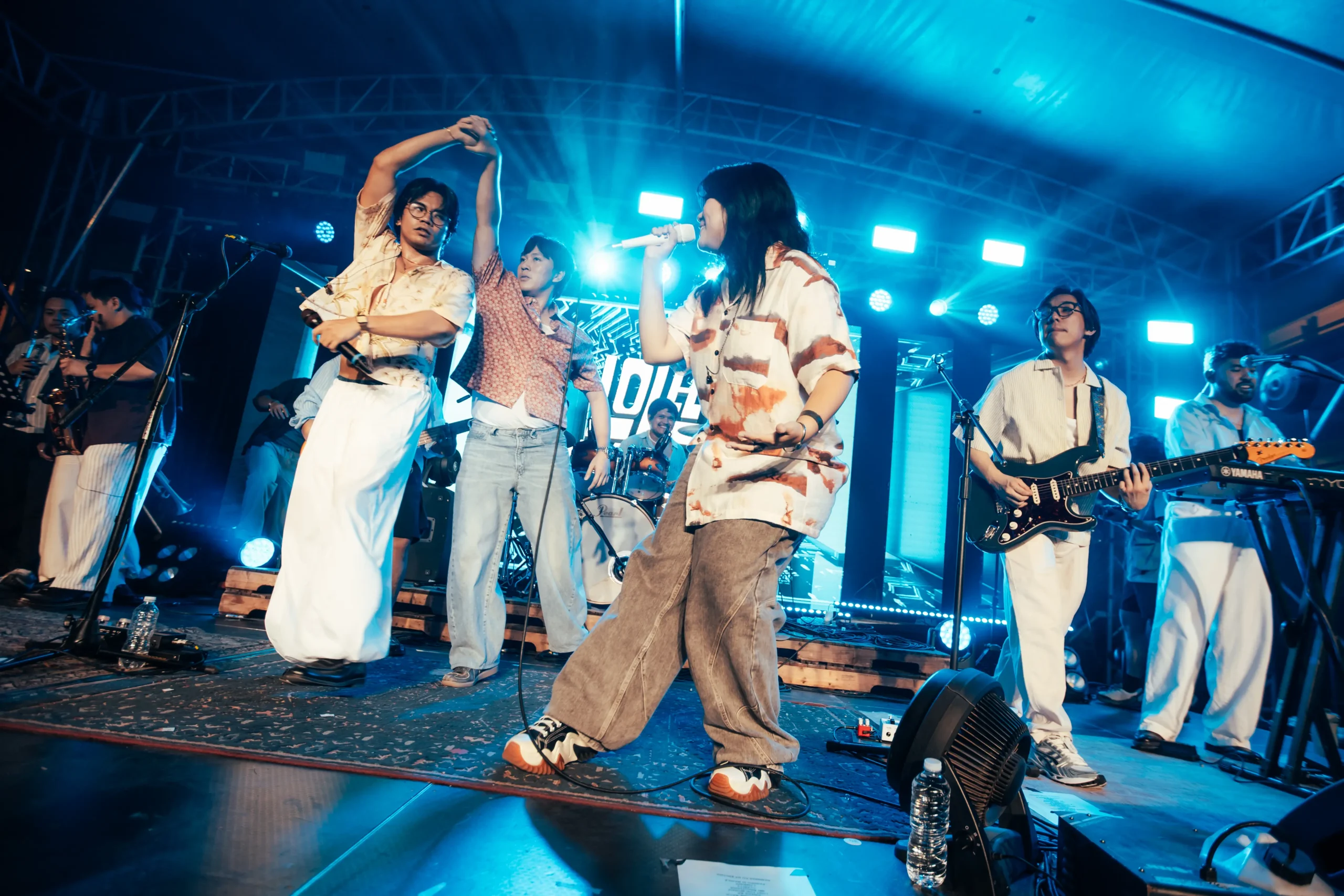 Band performing on a blue-lit stage; singer center with microphone, guitarist to the right, and a saxophonist to the left.