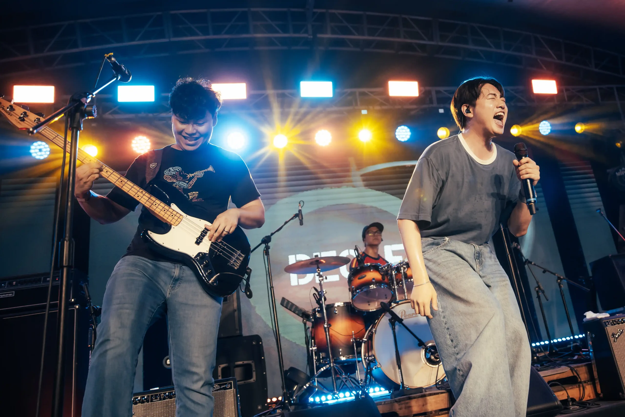 Two male musicians perform on a lit stage: a guitarist left and a vocalists right under colorful stage lights.