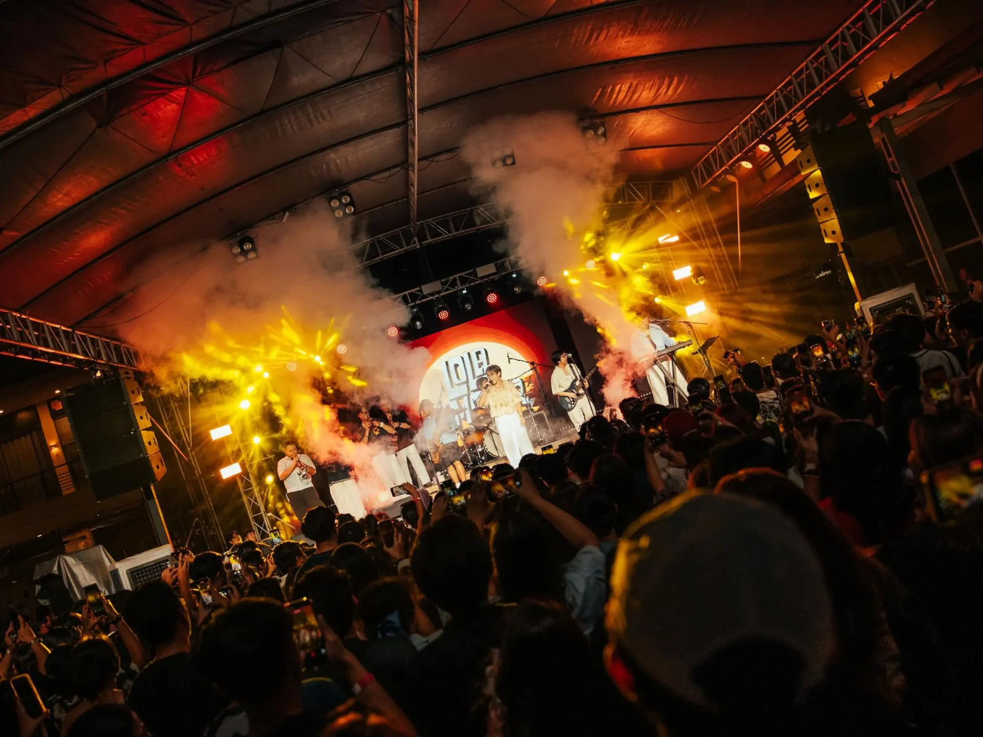 Concert stage with performers under a large tent, bright yellow stage lights and smoke, crowd cheering and recording with phones in the foreground