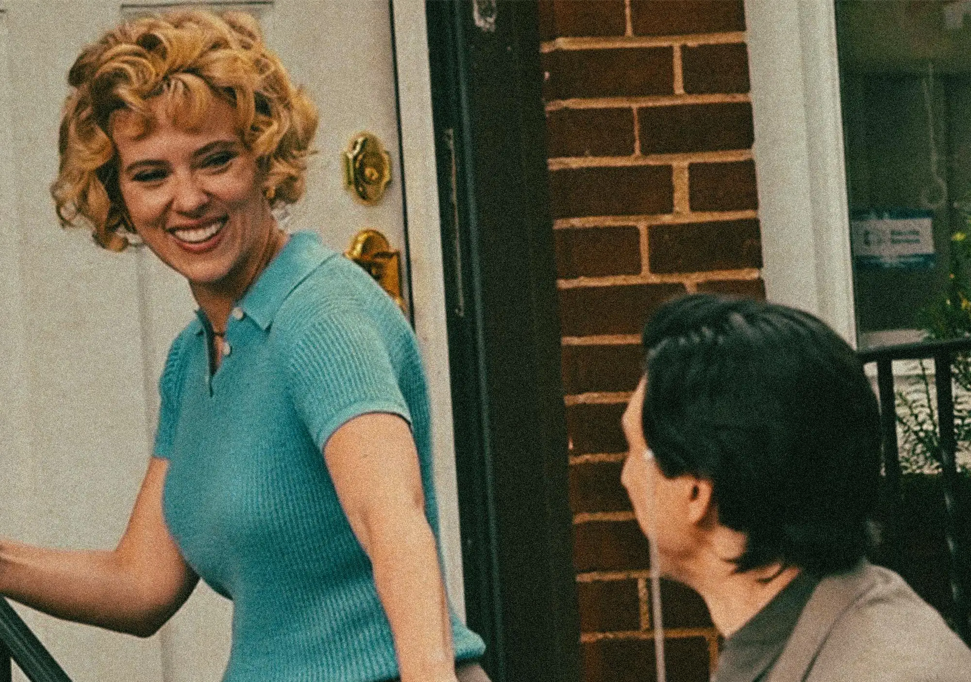 Smiling blonde woman in a teal shirt steps out of a doorway, with a brick wall outside and a child visible in the foreground.
