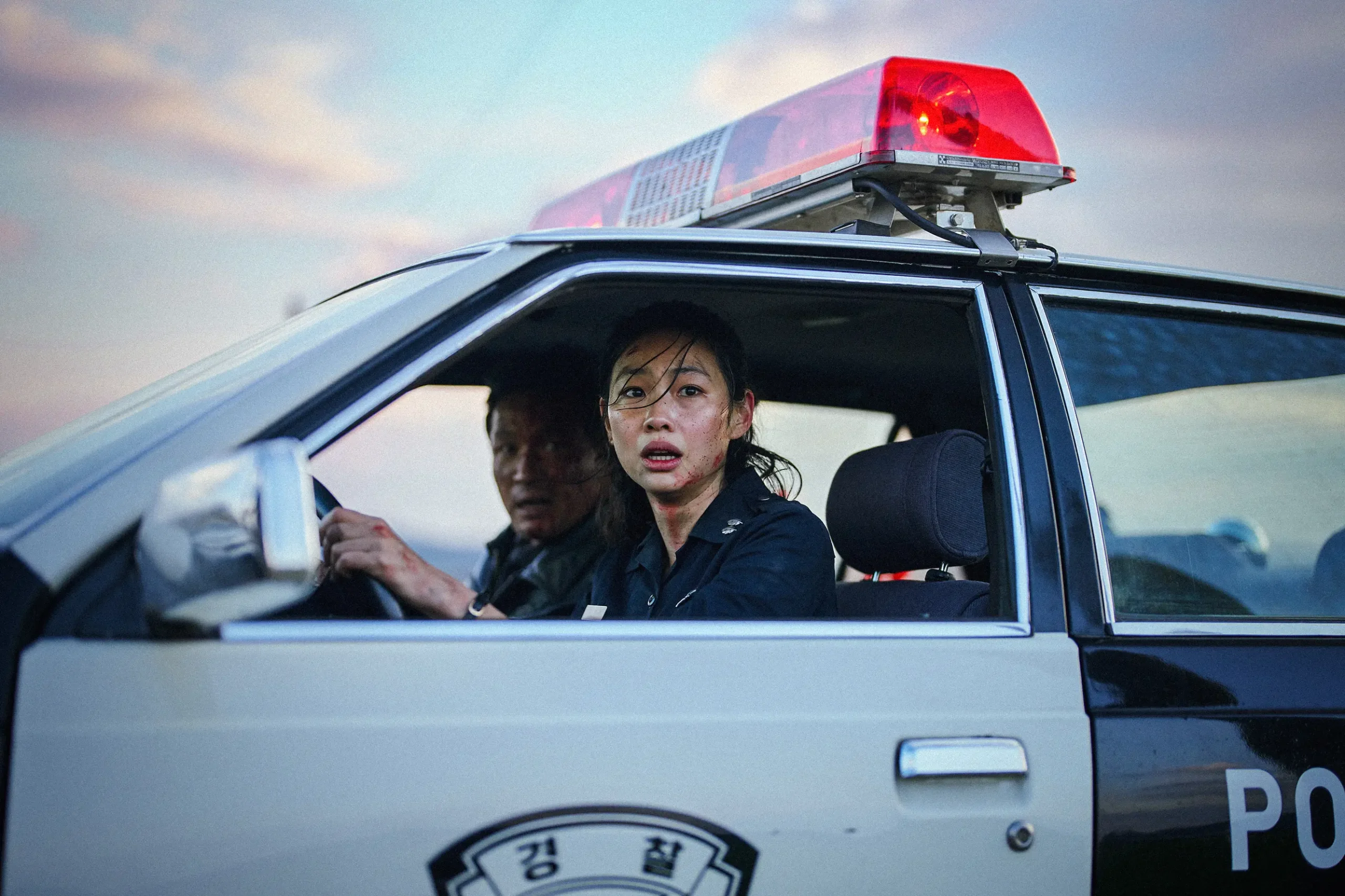 Police car with a red emergency light on top; two officers visible in the front seats looking ahead, serious expressions.