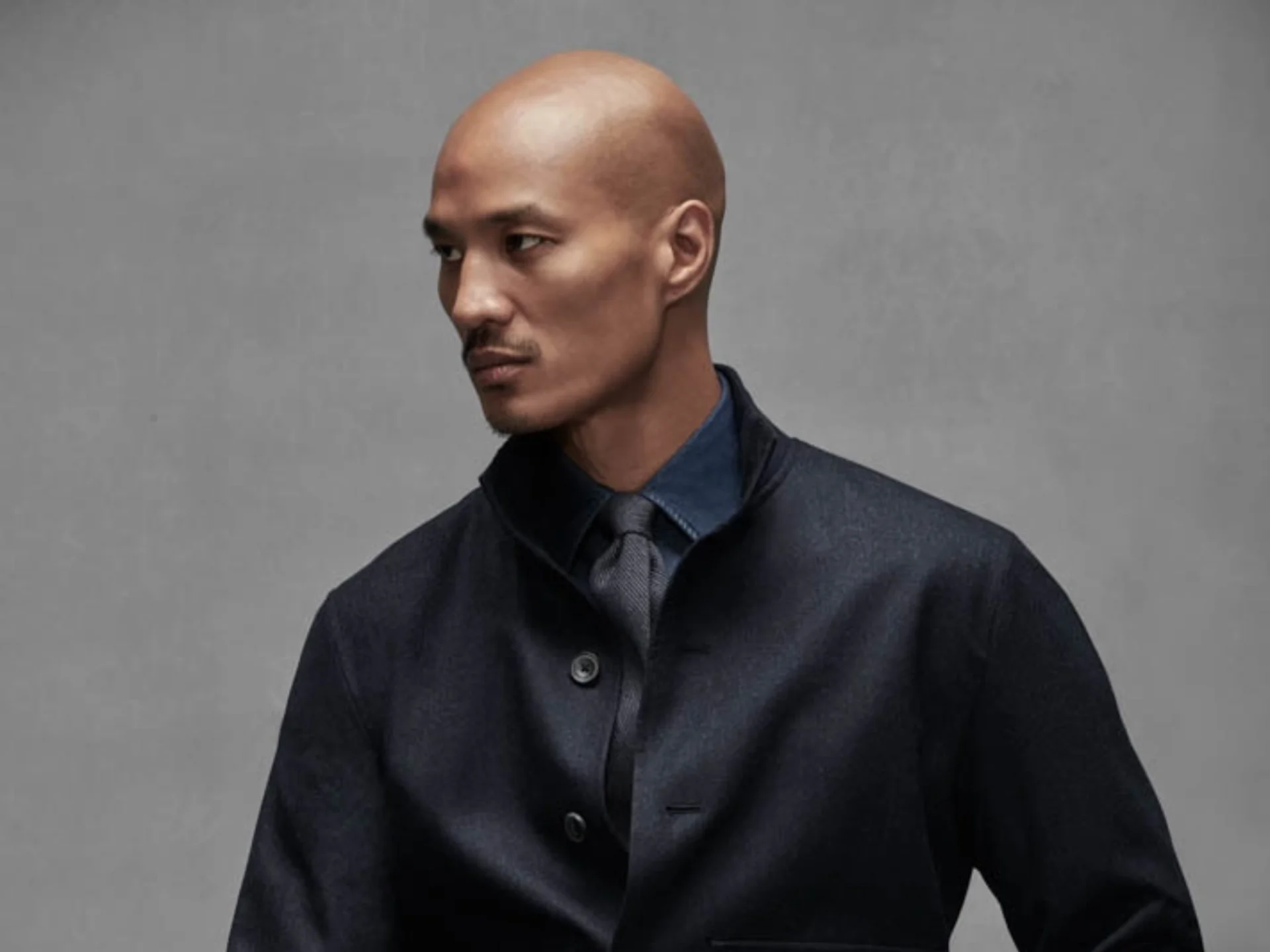 Bald man in a dark blazer and blue shirt turns to the left against a gray studio backdrop, serious expression.