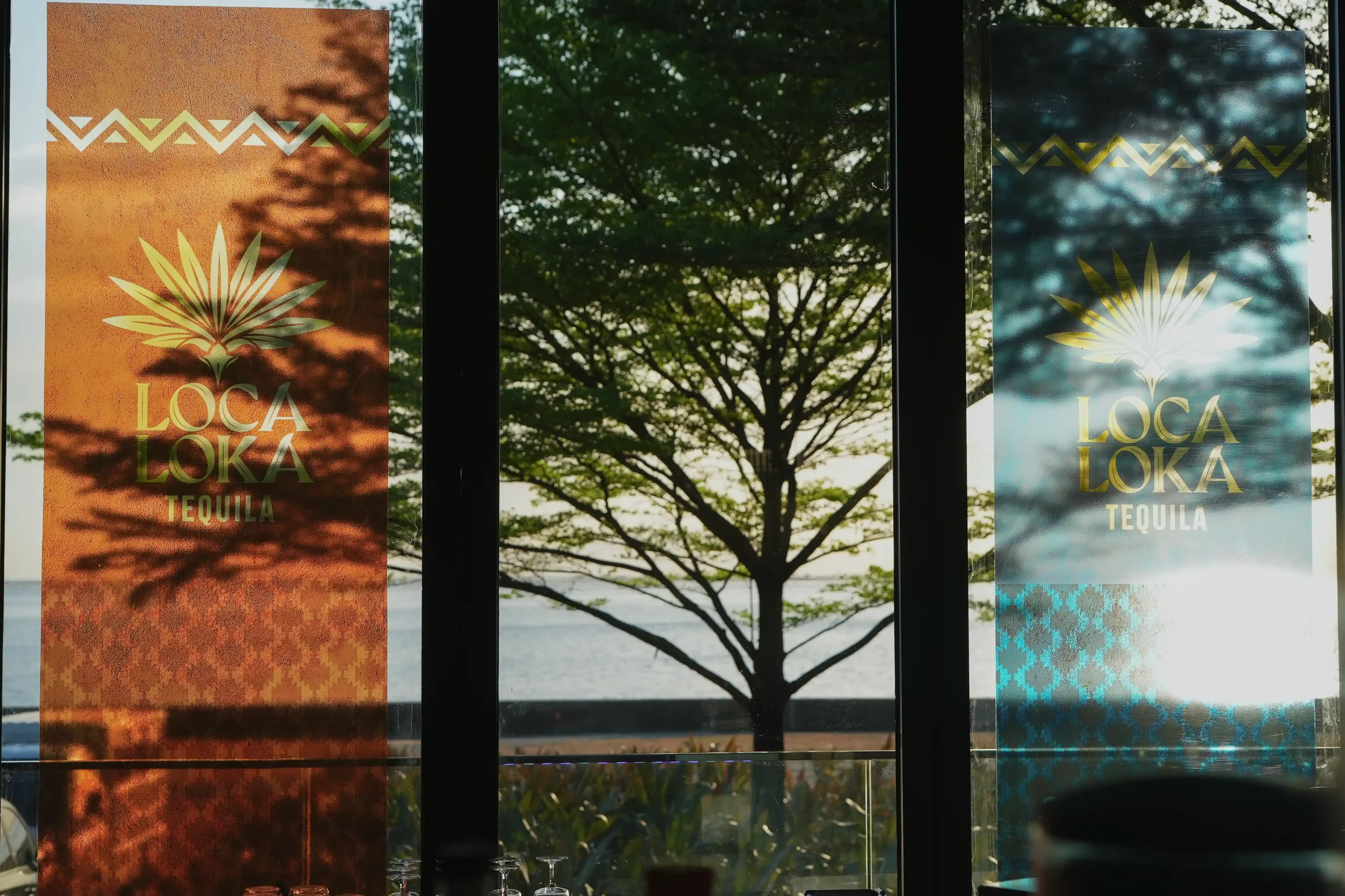 Two vertical posters on a glass storefront advertising LOCA LOKA Tequila, orange left panel and teal right panel with a tree reflected in the window.