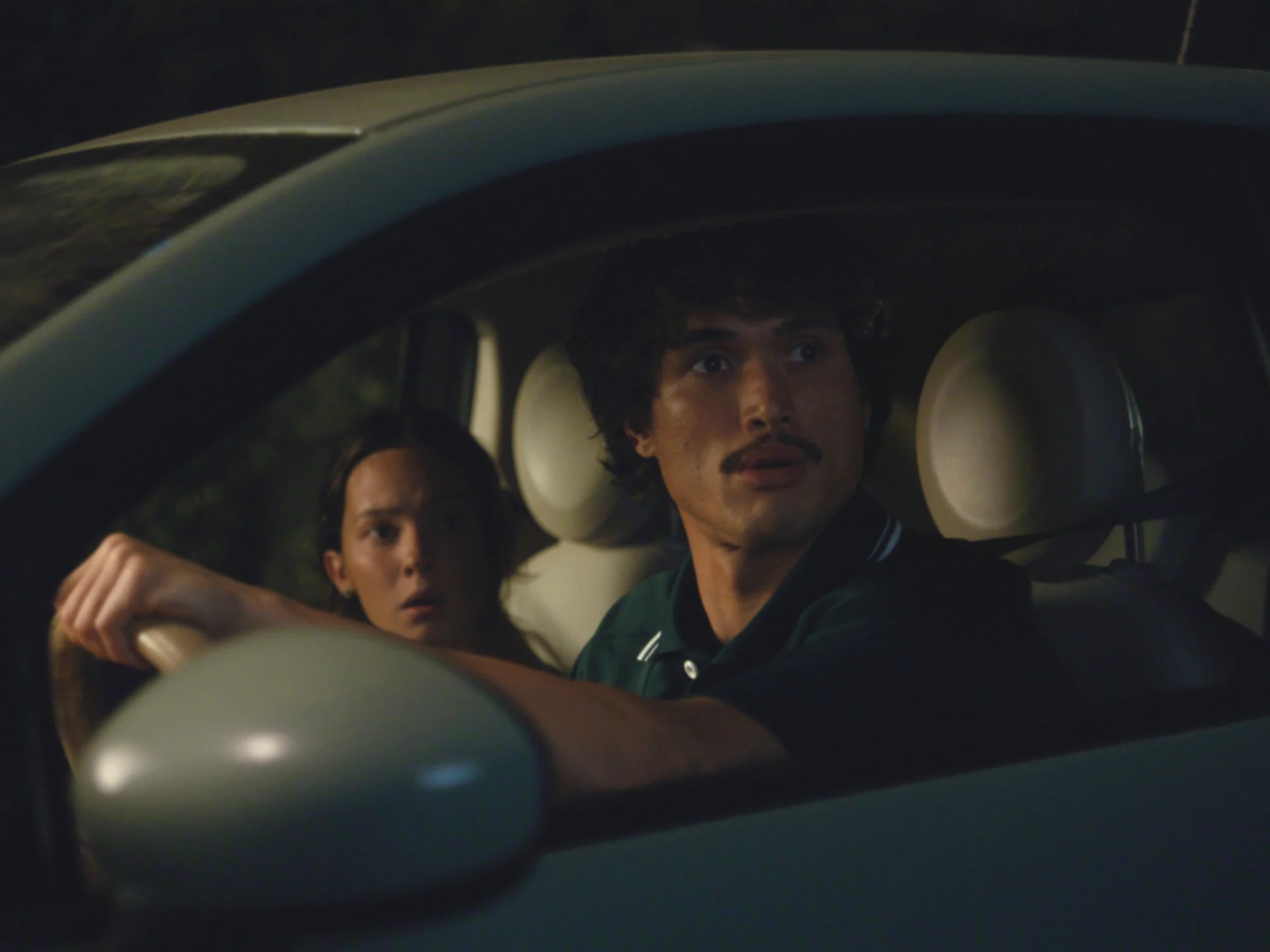 Two people inside a car at night, the man driving and the woman in the passenger seat, both looking alarmed.