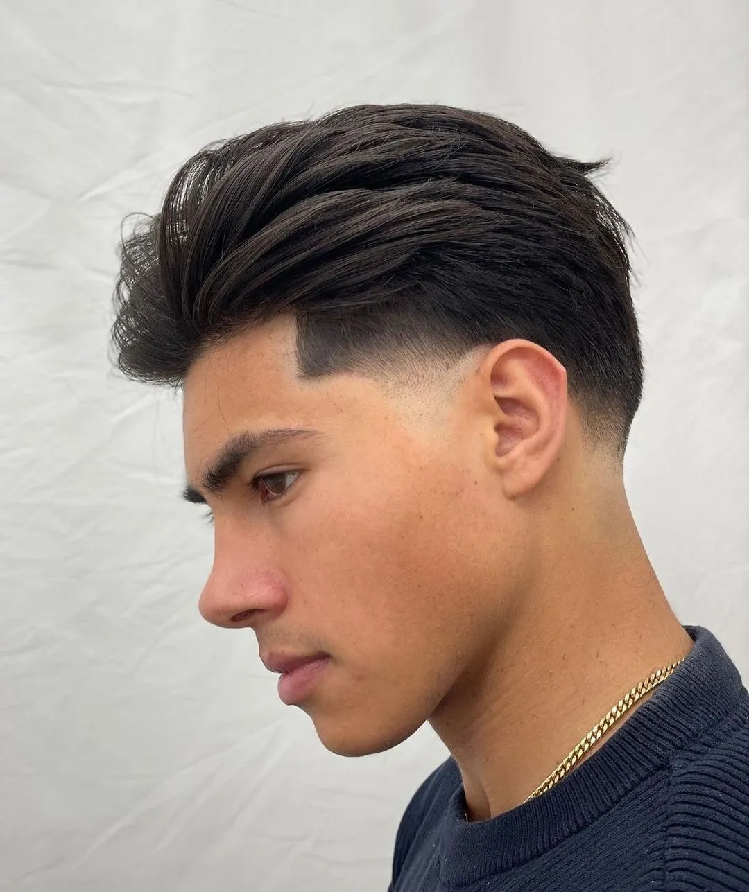 Profile view of a young man with a high fade haircut, dark slicked-back hair, wearing a dark sweater and gold chain against a light backdrop.