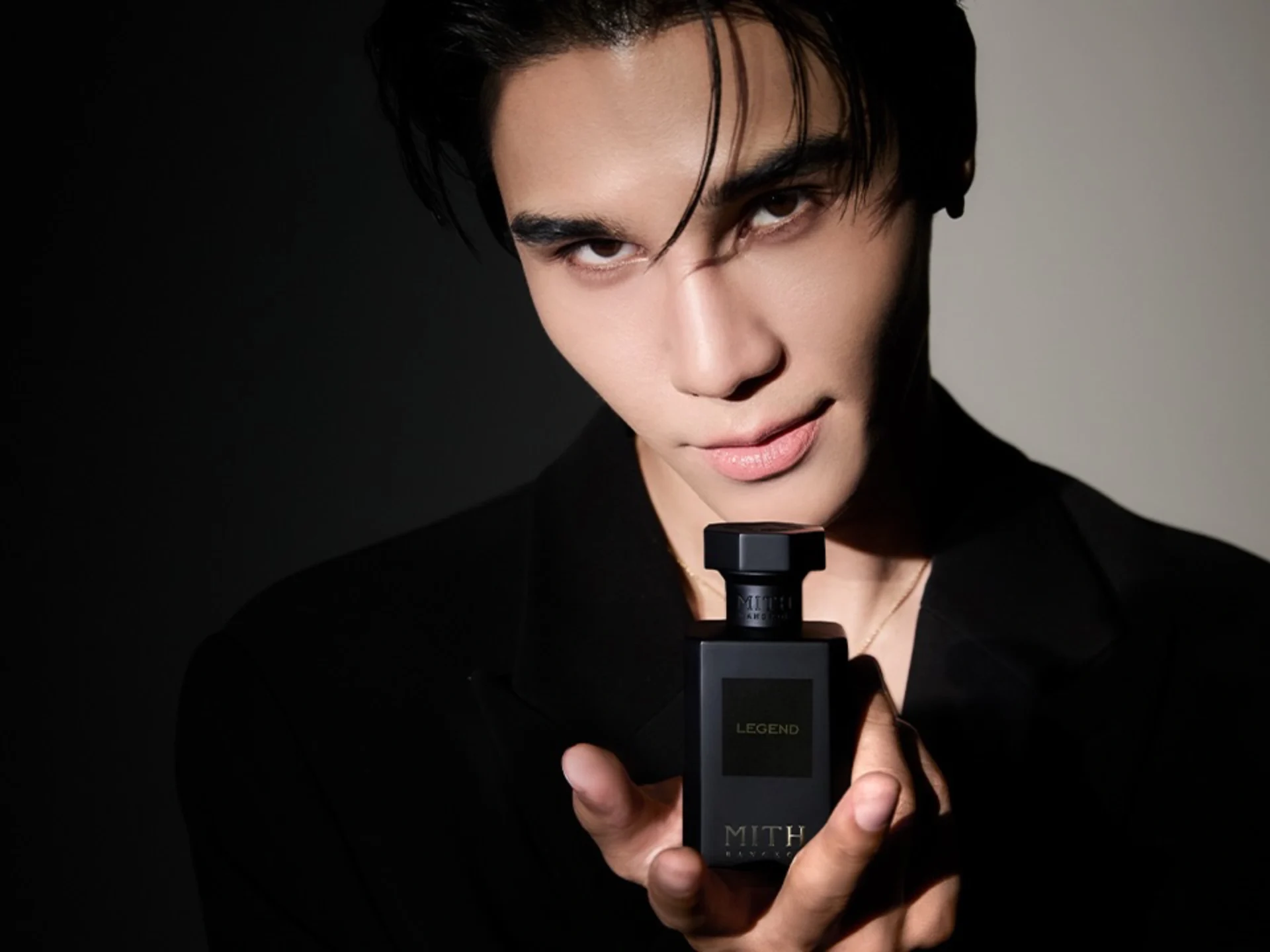 Close-up of a model's face with dark hair, holding a tall rectangular perfume bottle near her lips against a dark background.