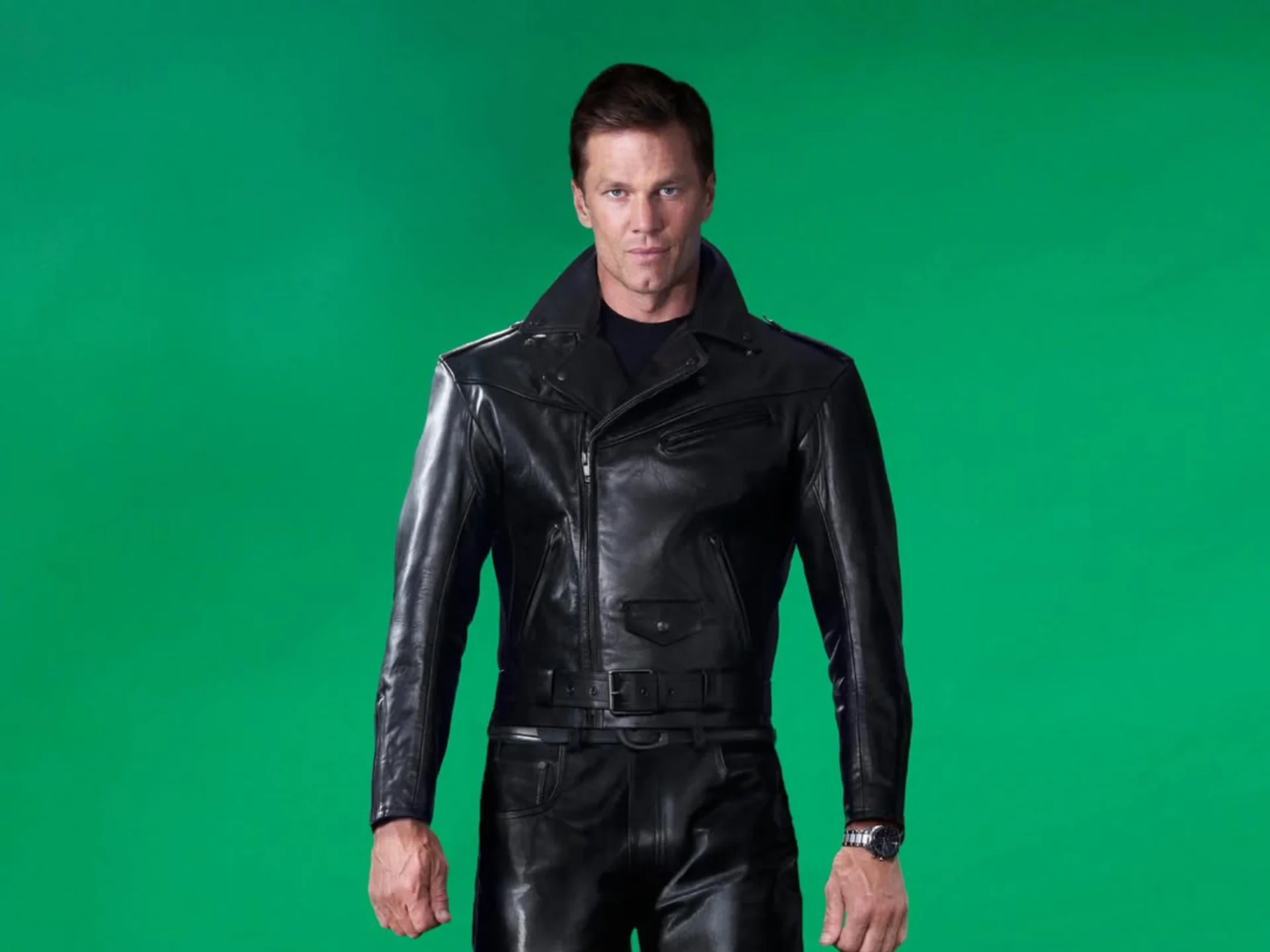 Man in a black leather jacket posing against a solid green background, looking at the camera.