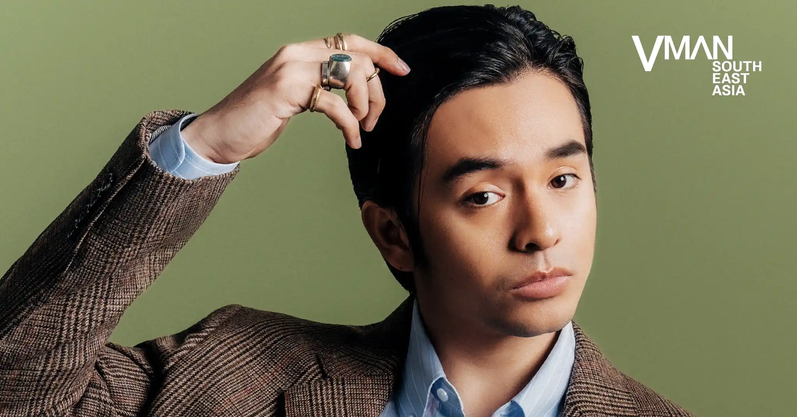 Person adjusting hair in a brown plaid blazer over a light blue shirt, against a green background with a VMAN South East Asia logo in the top right.