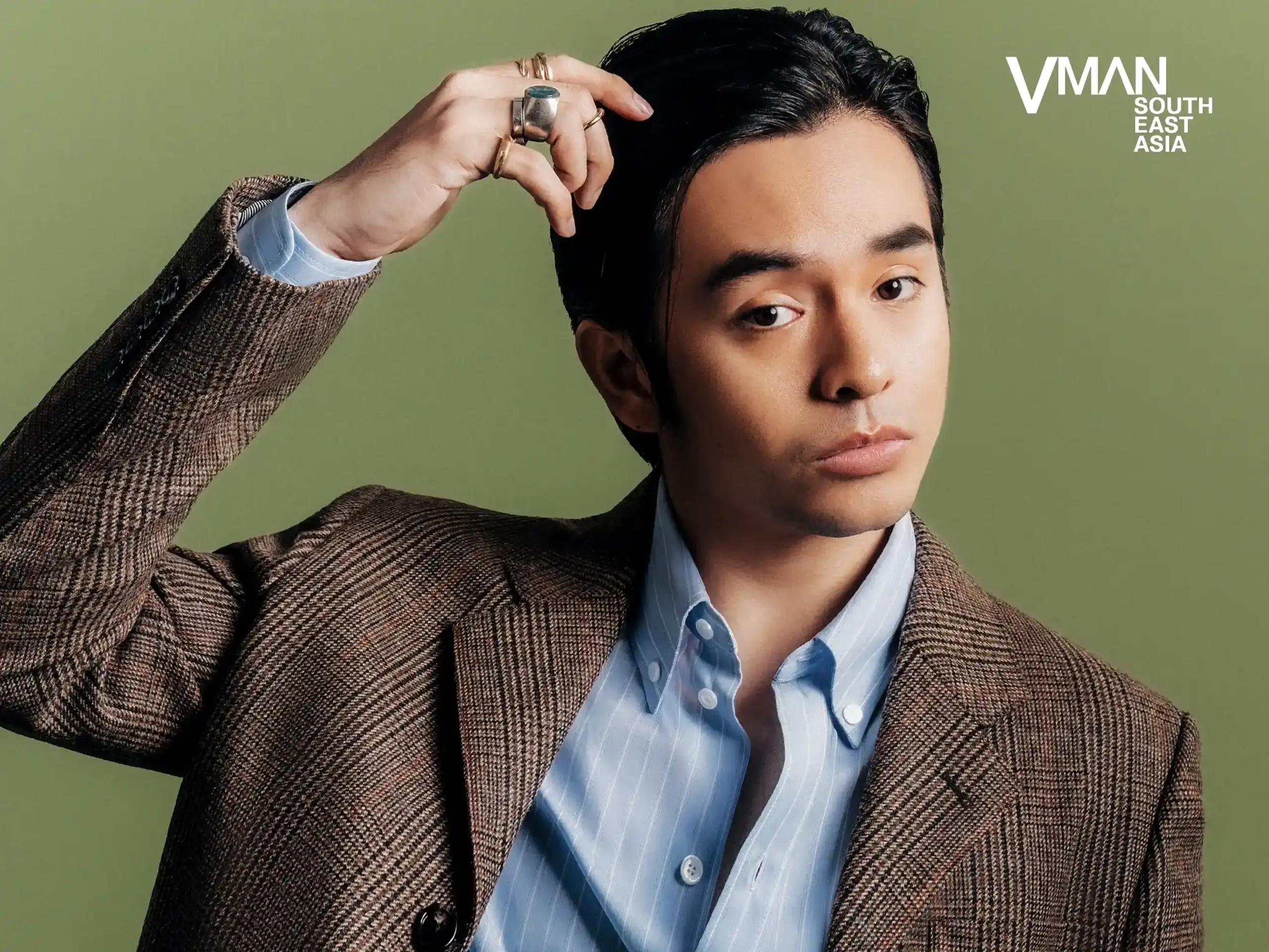 Person adjusting hair in a brown plaid blazer over a light blue shirt, against a green background with a VMAN South East Asia logo in the top right.