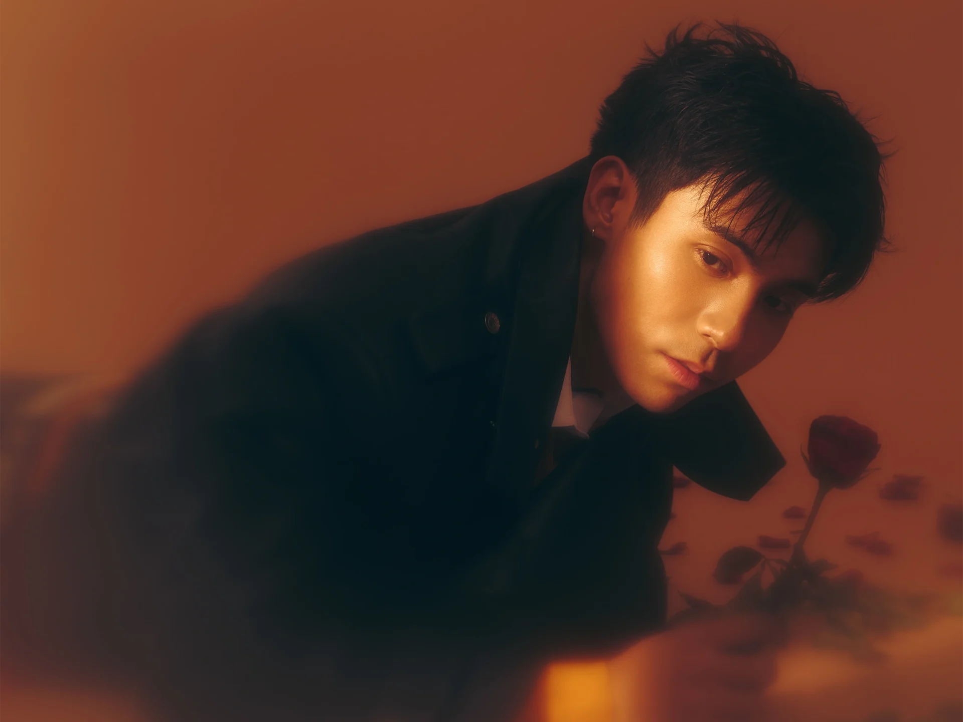 Close-up portrait of a person with short hair, wearing a dark jacket, bathed in warm orange light with roses in the background, looking at the camera.