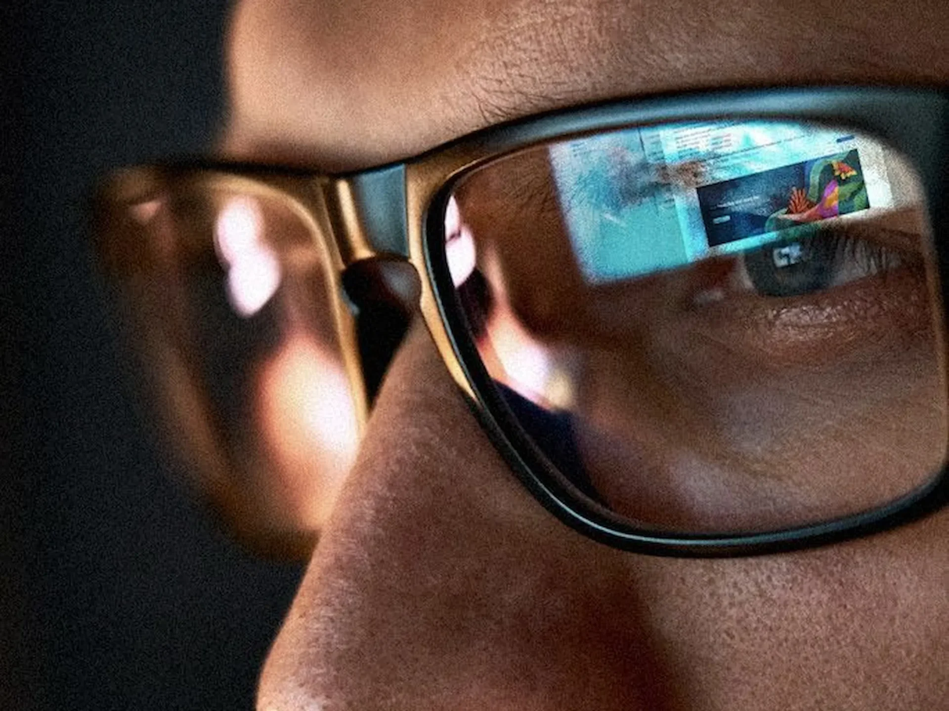 Close-up of a person wearing black-rimmed glasses; the right lens reflects a colorful digital screen.