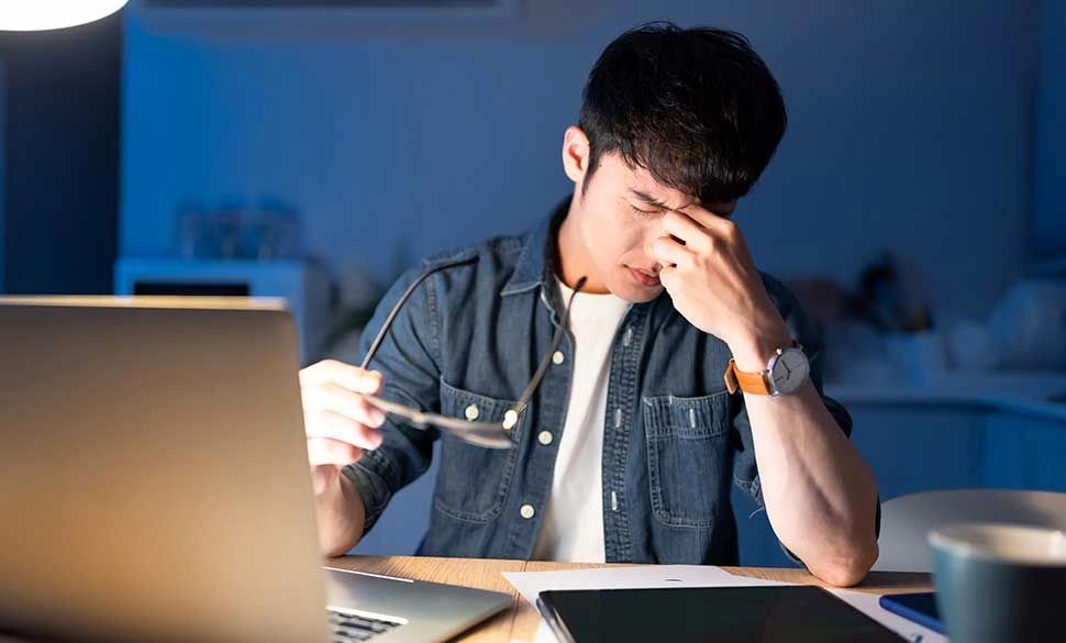 Man in a denim shirt at a desk, pinching the bridge of his nose while holding glasses and staring at a laptop in a blue-lit room.