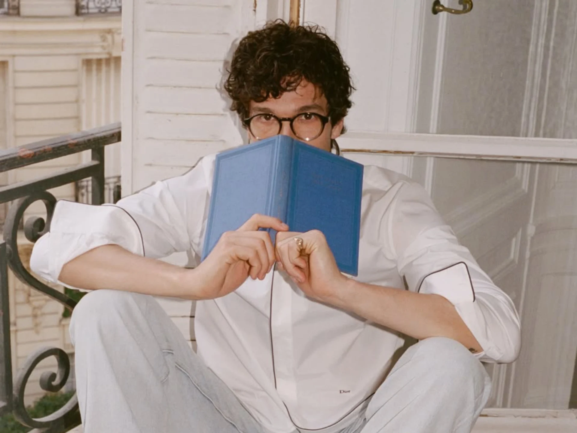 Person with curly hair and round glasses reads a blue hardcover book on a porch.