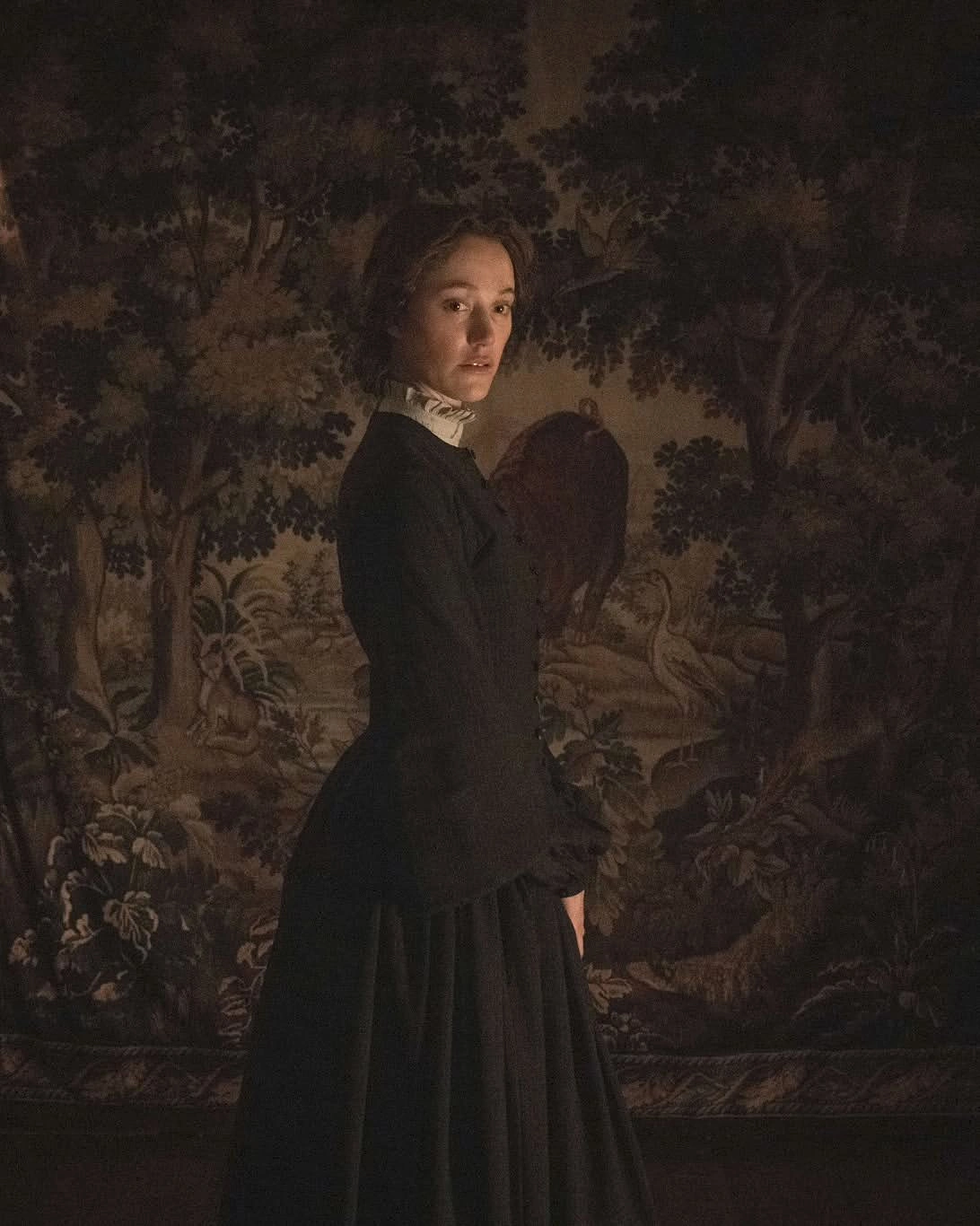 Woman in a dark Victorian-style dress with a white high collar, standing in a dimly lit room against a forest mural backdrop.