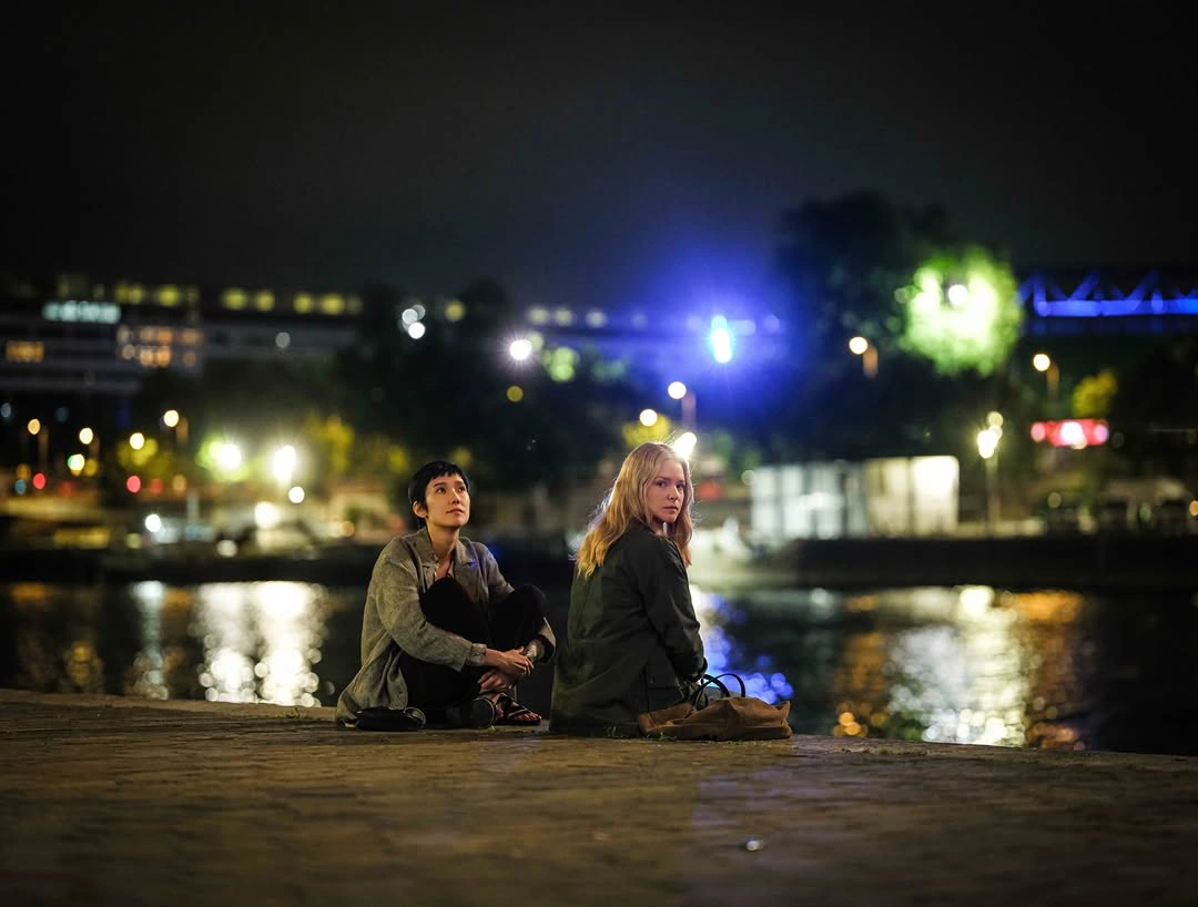 Two women sit on a waterfront boardwalk at night, gazing toward the city lights reflected on the water.