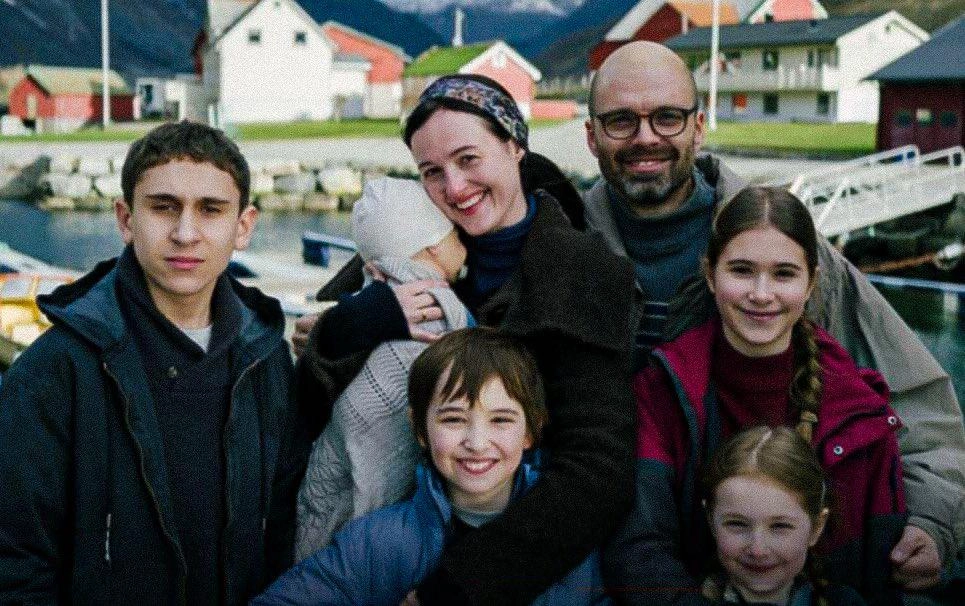 A smiling family of six posing outdoors by a harbor with colorful houses in the background.